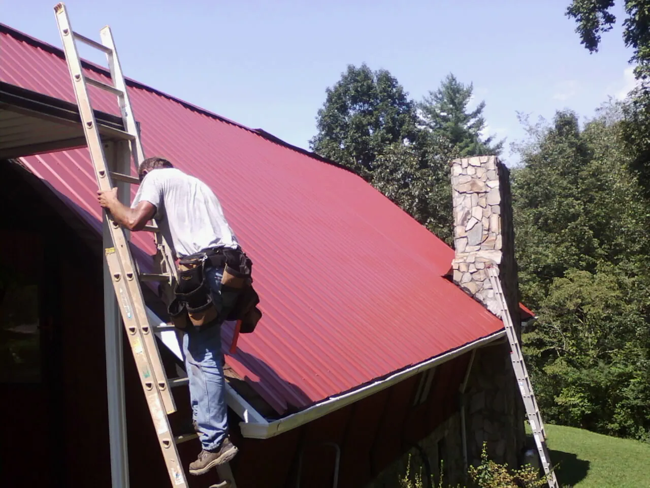 Roofer on a ladder beside a red metal roof and stone chimney — Soffit & Fascia Repair by Roofing Brothers in Udall